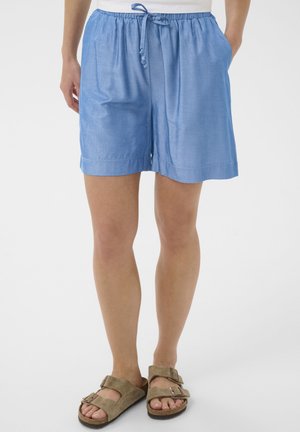 Femeie purtând pantaloni scurți bleu deschis cu șnur și buzunare, precum și sandale bej cu cataramă, stând în picioare pe un fundal alb simplu.