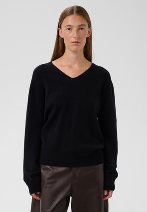 Giovane donna con capelli castani lisci che indossa un maglione nero a scollo a V e pantaloni in pelle marrone scuro, in piedi davanti a uno sfondo grigio semplice.