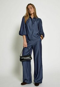 Chemise bleu marine à manches bouffantes et se fermant par des boutons, associée à un pantalon large. Le modèle tient un petit sac à main noir en cuir verni. Texture lisse.