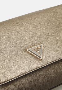 Kabelka s texturou zlaté barvy má trojúhelníkové kovové logo s názvem značky. Strukturovaný design a hladký vnější povrch.