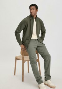 Green zip-up jacket over a cream shirt, paired with green pants and light sneakers. Model seated on a wooden chair against a neutral background.