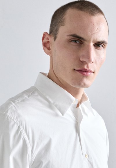 Camisa blanca de cuello, hecha de una tela de algodón suave, con un diseño clásico de botones y una textura ligeramente arrugada.