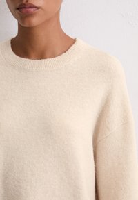 Pull en maille beige avec un col rond, texture douce et épaules tombantes. Le tissu semble chaud et confortable, avec des variations de couleur subtiles.