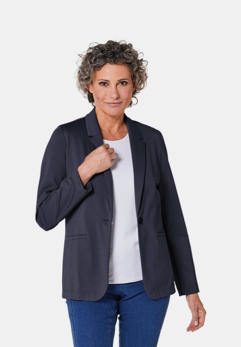 Donkerblauwe blazer met volledig omgeslagen revers, knoopsluiting, twee zakken en een gladde textuur, gecombineerd met een witte top en blauwe spijkerbroek.