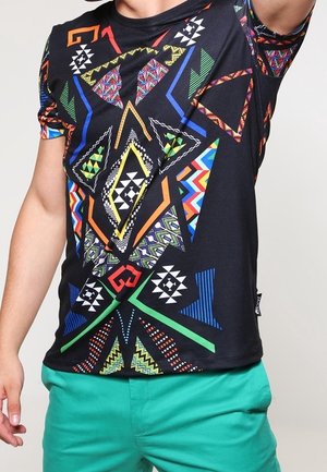 Czarny T-shirt z kolorowymi geometrycznymi i plemiennymi wzorami, noszony z zielonymi szortami na osobie z podniesioną ręką.