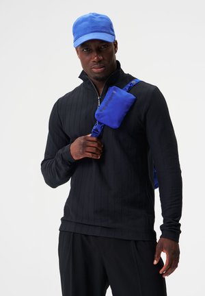 Schwarzes Langarmshirt mit gerippter Textur, blaue Kappe und eine blaue Umhängetasche mit einem Logo, die über der Schulter getragen wird.