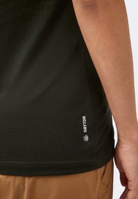 Κοντινό πλάνο του πλευρού ενός ατόμου που φοράει μαύρο t-shirt Dryton και καφέ παντελόνι, εμφανίζοντας μέρος του χεριού και του κάτω κορμού.