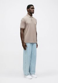 Béžové polo tričko s límečkem a krátkými rukávy, kombinované s světle modrými širokými džínami, nošené s bílými teniskami.