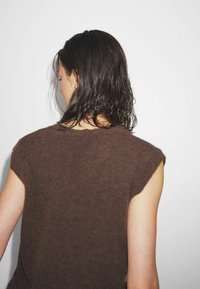 Brūns adīts bez piedurknēm tops ar apaļu apkakli. Mīksta tekstūra un brīvs piegriezums, parādot neapstrādātas malas pie rokas caurumiem. Mati maigi krīt ap pleciem.