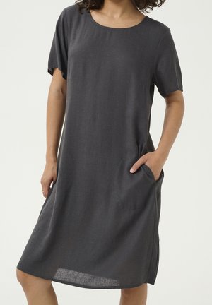 Femme portant une robe courte simple gris foncé avec des poches, debout devant un fond clair uni.