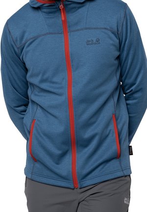 Personne portant une veste bleue zippée Jack Wolfskin avec une fermeture éclair rouge et des bordures de poches rouges, ainsi qu’un pantalon gris avec le logo Jack Wolfskin.