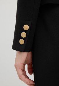 Manica di blazer in tessuto nero con tre bottoni dorati texture e un design elegante e su misura. Vista ravvicinata del dettaglio della manica.
