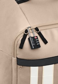 Beige rugzak van een gestructureerde stof, voorzien van een rits en een zwart cijferslot. Geaccentueerd met een contrasterende zwarte streep en witte lijnen.