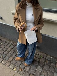 Hnědý vlněný kabát, bílý žebrovaný rolák, modré džíny se širokými nohavicemi, hnědé kotníkové boty, drží pečivo a bílou papírovou tašku. Kamenitá dlažba.