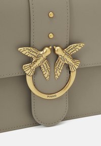 Pelēka ādas rokassoma ar zelta apļveida sprādzi, kurai ir divi detalizēti putnu akcenti un trīs zelta kniedes virsū. Redzams zīmola nosaukums "PINKO".