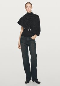 Kvinna klädd i en svart asymmetrisk stickad poncho, mörka raka jeans, svarta skor och ett långt hänge, stående mot en neutral bakgrund.