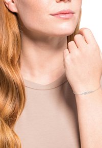 Kvinna med långt rött hår som bär en beige topp och ett delikat silverarmband, håller handleden nära ansiktet mot en vit bakgrund.