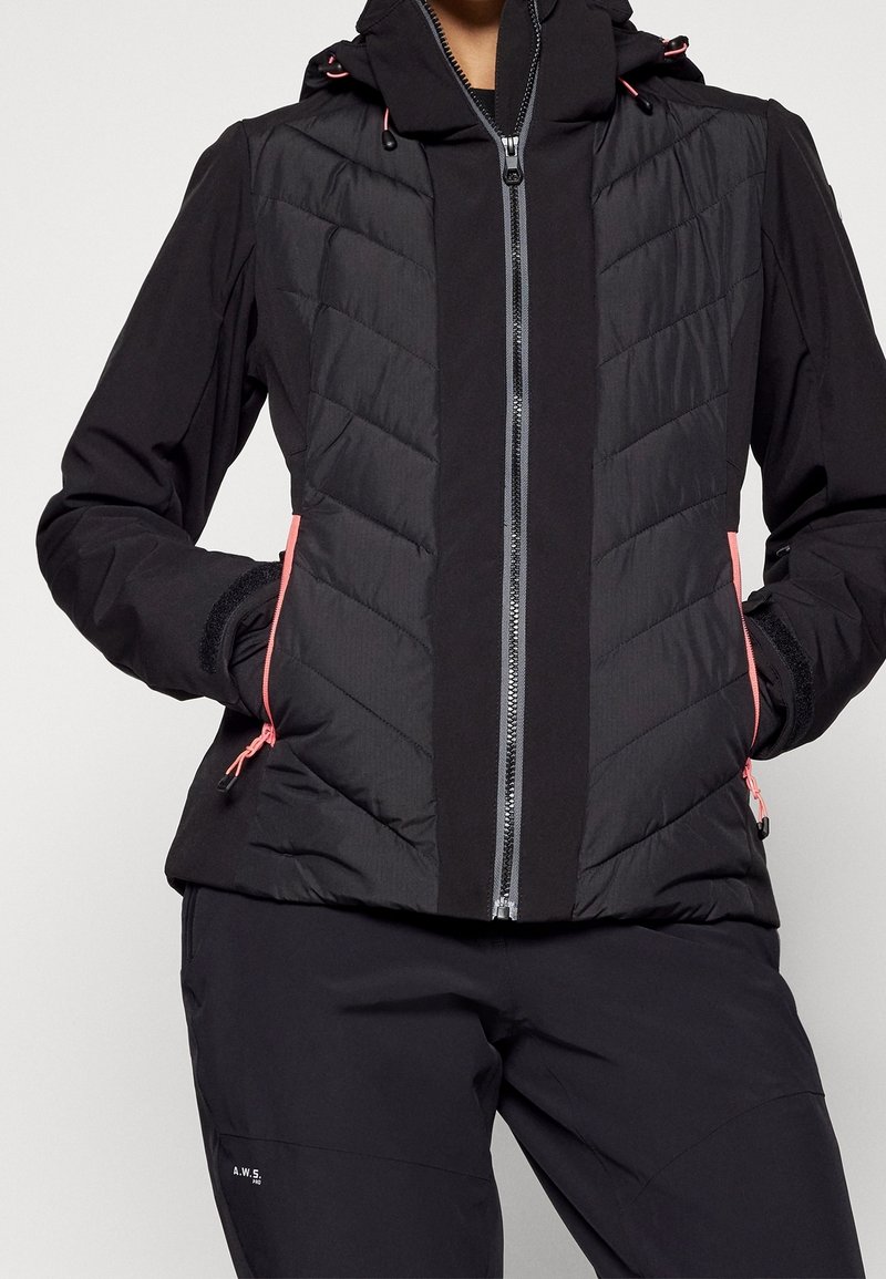 Personne portant une veste matelassée noire avec une fermeture éclair avant et des détails de fermeture éclair roses, associée à un pantalon noir, les mains dans les poches de la veste.