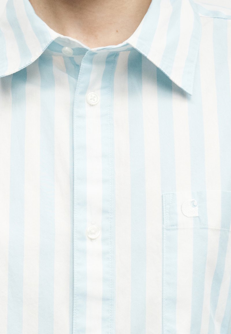 Primo piano di una persona che indossa una camicia a righe verticali azzurre e bianche, con colletto e piccoli bottoni, e una piccola tasca sul petto.