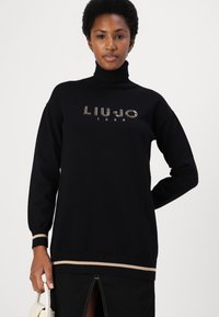 Zwarte coltrui met "LIUJO 1995" in strass, geribde manchetten en een contrasterende beige streep aan de onderkant. Zachte textuur.