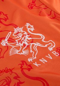 Oranje stof met een geborduurd wit leeuwenlogo dat een zwaard vasthoudt, met rode leeuwpatronen op de achtergrond. Eenvoudig en levendig ontwerp.