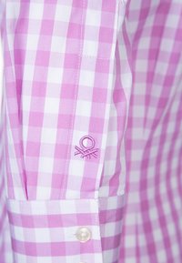 Rosa och vitrutig skjorta med knäppning vid ärmen som har en broderad logotyp i rosa. Slät bomullstyg med lätt textur.