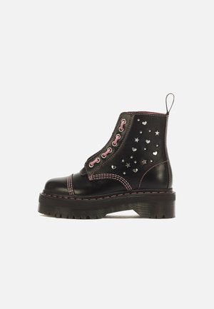 Bottine en cuir noir avec lacets roses, coutures roses et clous argentés en forme de cœur et d'étoile. Semelle épaisse à rainures et languette à l’arrière.
