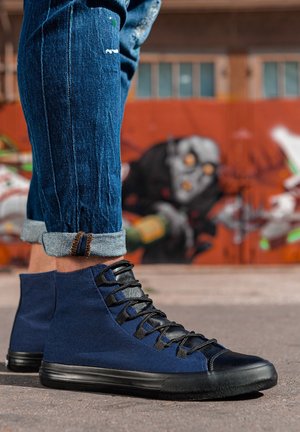Personne portant un jean bleu retroussé et des baskets montantes bleu marine, debout sur une surface pavée avec des graffitis sur un mur en arrière-plan.