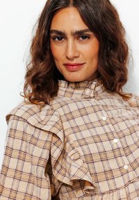Blouse à carreaux beige avec un col haut, des détails en volants sur les épaules et des boutons blancs. Présente un tissu texturé avec un motif à carreaux.