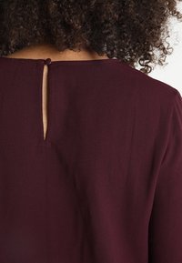 Haut bordeaux avec un détail dos en forme de trou de serrure, présentant une texture de tissu lisse et des manches courtes. Encolure finement cousue.