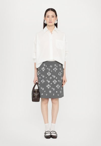 Hvitt oversized skjorte med knapper kombinert med et grått blyantskjørt som har blomstermotiver. Tilbehøret inkluderer en liten brun veske og hvite sokker.