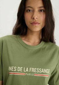 T-shirt en coton vert olive à manches courtes et col rond, avec un texte blanc et rouge indiquant "INES DE LA FRESSANGE PARIS."