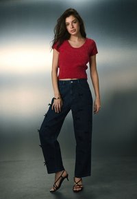 Crvena teksturirana majica kratkih rukava uparena s tamnoplavim trapericama ukrašenim crnim mašnama i crnim sandalama na remenčiće.