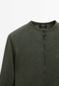 Donkergroene linnen cardigan met een ronde hals, knoopsluiting aan de voorkant en een gestructureerde afwerking. Voorzien van vijf zwarte knopen langs de knoopsluiting.