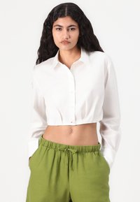 Camisa blanca corta con cierre de botones y mangas largas, combinada con pantalones verdes con cordón ajustable. Material de algodón, textura suave, diseño sencillo.