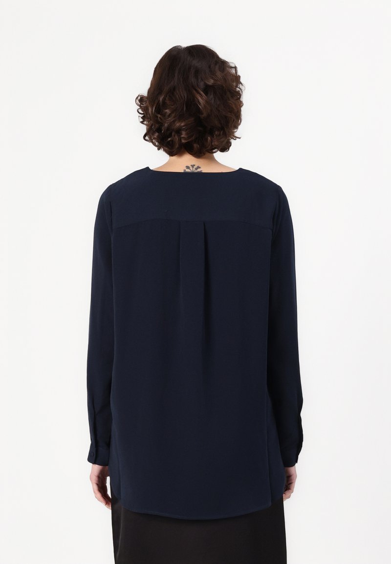 Marineblaue Bluse mit langen Ärmeln und rundem Halsausschnitt, Falten-Detail auf dem Rücken und geknöpften Manschetten. Der Stoff wirkt glatt und leicht.