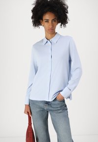 Woman with curly hair wearing a light blue button-up shirt and jeans, holding a red bag in one hand and the other hand in her pocket.