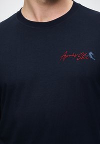 Granatowa bawełniana koszulka z okrągłym dekoltem, z haftowanym na czerwono i niebiesko napisem "Apres Ski" oraz sylwetką narciarza na lewej piersi.