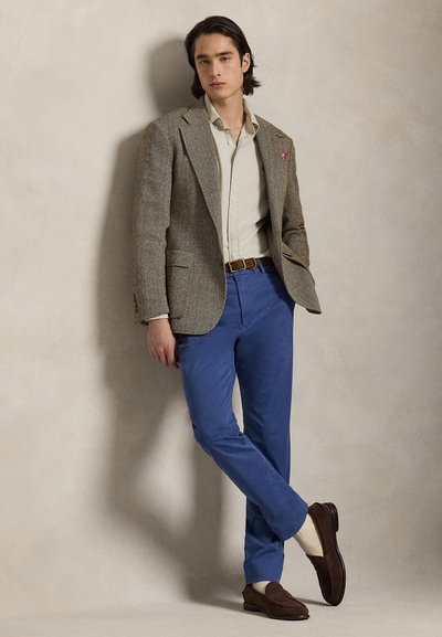 Καφέ σακάκι με σχέδιο ψαροκόκκαλο, ανοιχτό μπεζ πουκάμισο, ζωντανό μπλε παντελόνι, καφέ σουέντ μοκασίνια και κρεμ κάλτσες. Στάση ορθίας με ουδέτερο φόντο.