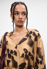 Καφέ μπλούζα με V-λαιμό, που διαθέτει αφηρημένο σχέδιο σε μαύρο και σκούρο καφέ χρώμα. Ελαφρύ ύφασμα με διακριτική γυαλάδα, σε συνδυασμό με μια μπορντό τσάντα.