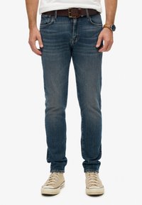 Hombre que lleva puestos jeans azules desgastados, un cinturón de cuero marrón, zapatillas blancas, un reloj de pulsera con correa de cuero marrón y una camisa blanca metida por dentro.