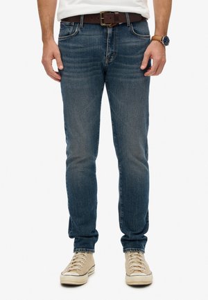 Man in vervaagde blauwe jeans, een bruine leren riem, witte sneakers, een bruine leren horloge met een leren band en een wit overhemd dat in zijn broek is gestoken.