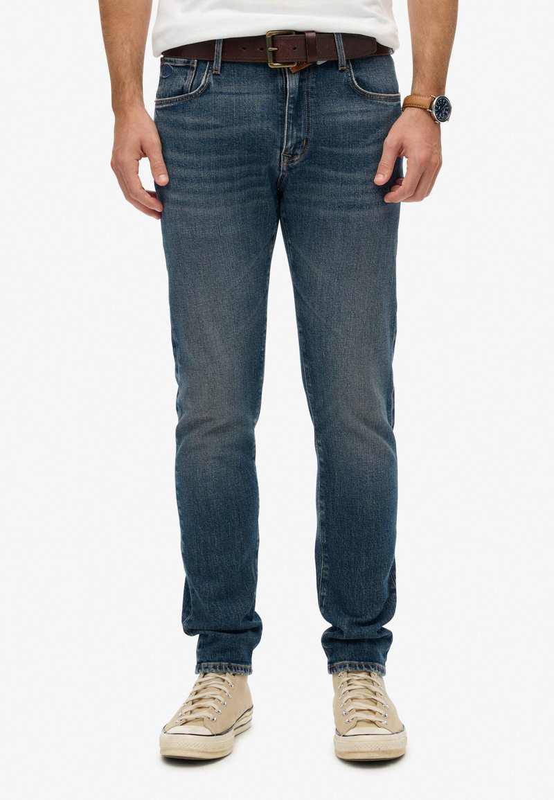 Hombre que lleva puestos jeans azules desgastados, un cinturón de cuero marrón, zapatillas blancas, un reloj de pulsera con correa de cuero marrón y una camisa blanca metida por dentro.