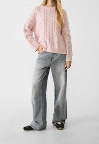Jasnoróżowy sweter w warkocze z okrągłym dekoltem, w połączeniu z szarymi dżinsami o wysokim stanie i szerokimi nogawkami oraz czarnymi sneakersami.