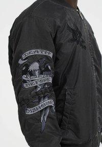 Black bomber jacket featuring embroidered eagle and a banner with "DEATH BEFORE DISHONOR" on the sleeve. Textured fabric and zip closure.