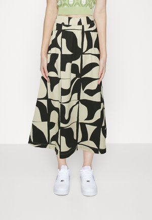Femme portant une jupe midi à motifs abstraits beige et noire, un pull court vert et des baskets blanches, debout devant un fond blanc uni.