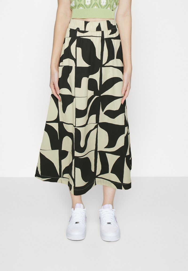 Femme portant une jupe midi à motifs abstraits beige et noire, un pull court vert et des baskets blanches, debout devant un fond blanc uni.