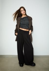 Bruine oversized jas, zwarte broek met wijde pijpen en grijs gestreept langärmelig shirt. Soepele texturen, relaxte pasvorm en minimalistisch ontwerp.