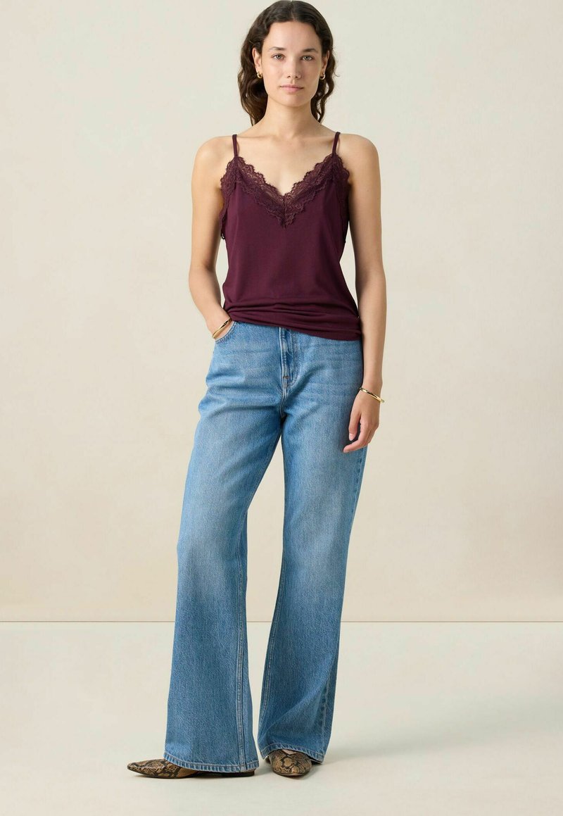 Vrouw staat in een mouwloos paars topje met kantafwerking, lichte blauwe wijde jeans en puntige schoenen met slangendessin.