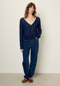 Granatowy sweter z dzianiny z głębokim dekoltem w serek i długimi rękawami, w zestawie z ciemnoniebieskimi dżinsami z wysokim stanem i brązowymi plecionymi sandałami.